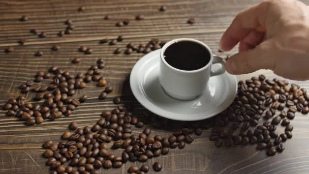 Mão masculina leva uma caneca de café. xícara de café e grãos de café. Xícara branca de café fumegante na mesa com feijão torrado . — Vídeo de Stock