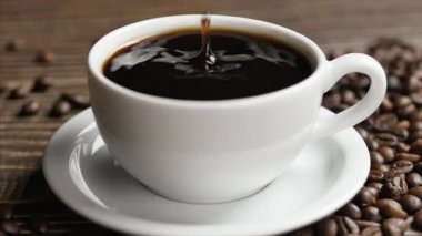 Tahta bir masanın üzerinde beyaz bir fincan kahve ve kahve çekirdekleri duruyor, yakın plan kahve damlaları bir fincanın içine düşüyor..