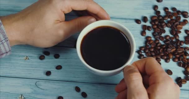 Mão masculina leva uma caneca de café. xícara de café e grãos de café. Xícara branca de café fumegante na mesa com feijão torrado . — Vídeo de Stock