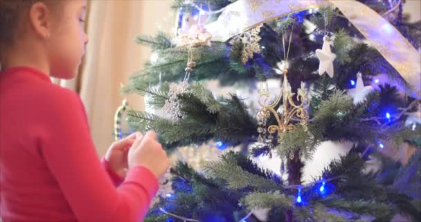 Klein meisje of kind hangt Nieuwjaarsspeelgoed aan de kerstboom, siert de kerstboom. Kerstversiering aan boom hangen met kerstverlichting. Decoreren op kerstboom met bal. 4k — Stockvideo