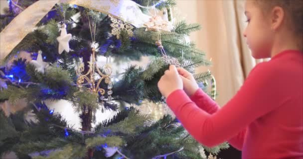 Маленька дівчинка або дитина вивішує іграшки Нового Року на ялинці, прикрашає ялинку. Повісити різдвяні прикраси на дереві з різдвяними вогнями. Декорація на ялинці з м'ячем. 4k — стокове відео