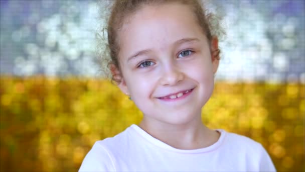 Retrato de uma menina pequena com olhos verdes olha para a câmera, contra um fundo de branco e ouro brilho brilhante. Retrato de um bebê engraçado ou criança sorrindo, olhando para a câmera . — Vídeo de Stock