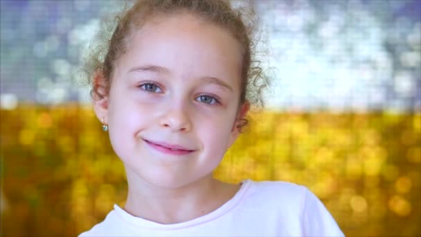 Portrait of a little young girl with green eyes looks at the camera, against a background of white and gold shiny glow. Portrait of a funny baby or child smiling, looking at camera. — Stockvideo