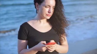 Mutlu, rahat, genç bir kadın ya da kız, deniz dalgalarının arka planında akıllı bir telefon tutuyor cep telefonu ekranına bakıyor, gülüyor, alışveriş için mobil uygulamalar kullanıyor, sosyal medyada sohbet ediyor..
