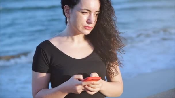 Happy relaxed young woman or girl holding smart phone on the background of sea waves looking at cellphone screen laughing enjoying using mobile apps for shopping having chatting in social media. — Stock Video