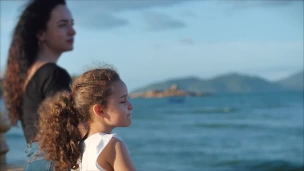 Anne ve kızından oluşan mutlu Avrupalı bir aile gün batımında sahilde durur ve okyanus ya da deniz kenarına bakar. Ağır çekim. Aile, tatil ve seyahat kavramı. — Stok video