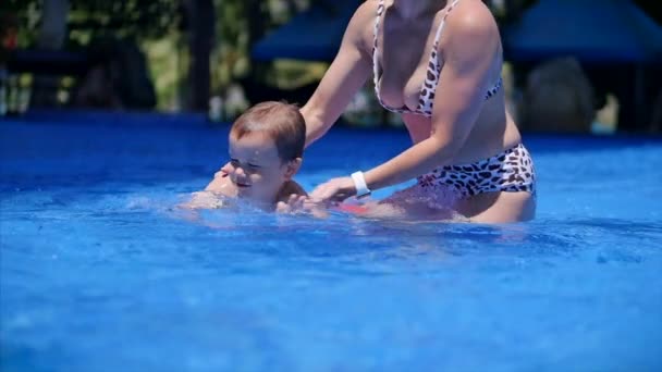 Mama albo opiekunka trzyma syna w ramionach, przytula go, uczy pływać. Mały chłopiec uśmiecha się szczęśliwie. Baby Boy pływać w basenie. — Wideo stockowe