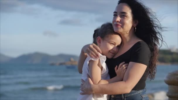 Familia feliz, madre y una linda hija en su suave abrazo se sientan en un banco cerca del mar, el niño con su amada madre sonríe y se abrazan firmemente. Concepto amor de padres e hijos — Vídeos de Stock