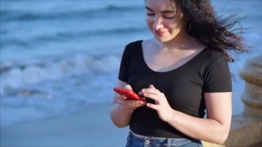 Mutlu, rahat, genç bir kadın ya da kız, deniz dalgalarının arka planında akıllı bir telefon tutuyor cep telefonu ekranına bakıyor, gülüyor, alışveriş için mobil uygulamalar kullanıyor, sosyal medyada sohbet ediyor..
