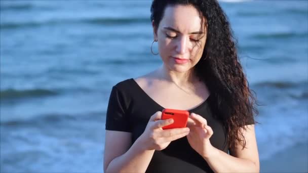 Mutlu, rahat, genç bir kadın ya da kız, deniz dalgalarının arka planında akıllı bir telefon tutuyor cep telefonu ekranına bakıyor, gülüyor, alışveriş için mobil uygulamalar kullanıyor, sosyal medyada sohbet ediyor.. — Stok video
