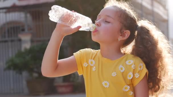 Schattig meisje lessen dorst, drinkt uit een plastic fles op straat in de buurt van het huis. Slow motion kleine meid drinkt gretig drinkwater. Close-up. — Stockvideo
