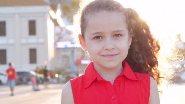 在一个阳光灿烂的夏日，一个滑稽的小女孩微笑着看着街上的摄像机，脸上挂着一个学龄前儿童的快乐的表情. — 图库视频影像