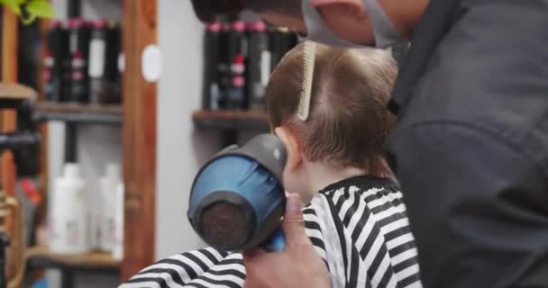 科罗纳韦病毒流行时，孩子们在沙龙里理发。小男孩理发了。孩子们的发型很时髦。穿着Covid-19型防护面罩的造型师使发型成为时尚的发型。 — 图库视频影像