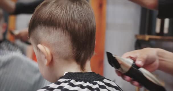 Childrens haircut in the salon at the time of the coronavirus epidemic. Little boy gets a haircut. Childrens hair styling. Stylist in a protective mask from covid-19 makes a stylish hairstyle for a — Stock Video