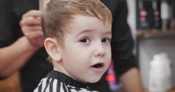 Coupe de cheveux d'enfants dans le salon au moment de l'épidémie de coronavirus. Le petit garçon se fait couper les cheveux. Coiffure d'enfant. Styliste dans un masque de protection de covid-19 fait une coiffure élégante pour un — Video