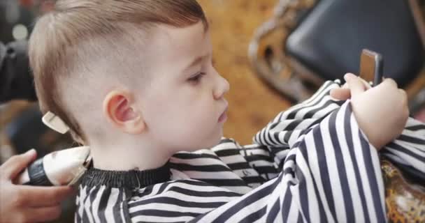 Little boy is watching a cartoon on a smartphone while the master makes his  haircut. Stylist in a protective mask from Covid-19 makes a stylish  hairstyle for a little boy. — Stock