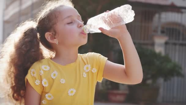 Şirin kız, evin yakınındaki sokakta plastik bir şişeden içiyor. Yavaş çekim küçük kız hevesle içme suyu içiyor. Yakın plan.. — Stok video