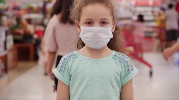 코로나 바이러스가 유행하는 유행병을 앓고 있는 어린이는 공포 속에서 쇼핑을 하고 있는 바이러스에 가려진 사람들 사이에서 카메라를 본다. — 비디오