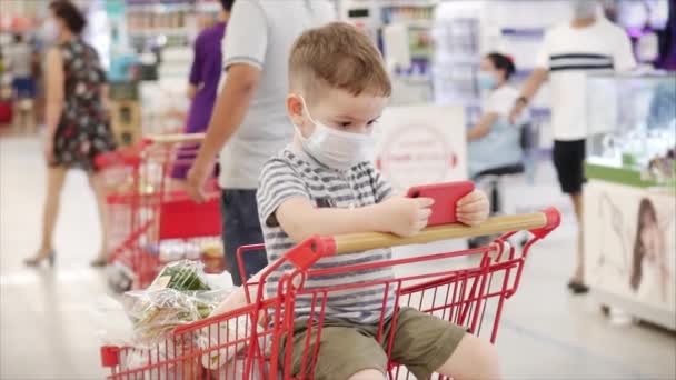 Familie kauft im Supermarkt ein, das Kind sitzt in einer Schutzmaske vor Viren, schaut Videos auf dem Smartphone.