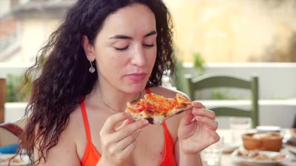 Mutlu, rahat, kırmızı elbiseli genç bir İtalyan kadın elinde pasta dilimiyle İtalyan pizzasının tadını çıkarırken aynı zamanda gülümsemesini seyrediyor ve evde lezzetli bir hamur işi yiyor. — Stok video