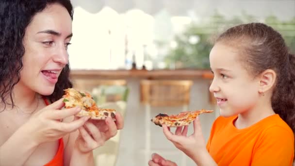 Linda menina filha com cabelo encaracolado senta-se com sua mãe na mesa comendo pizza italiana, olhando um para o outro sorrindo em resposta . — Vídeo de Stock