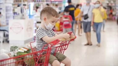 Çocuklu anne hipermarkette alışveriş yapıyor, çocuk virüslere karşı koruyucu bir maske takıyor, çocuk market arabasında oturuyor, videolar izliyor, telefonda çizgi film izliyor. Kavramsal İnsanlar, Koronavirüs, Salgın.