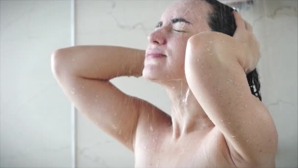 Mujer bastante joven lava el champú de su cabello mientras está de pie bajo un arroyo de ducha caliente. Concepto de limpieza del cabello y del cuerpo, higiene. — Vídeo de stock