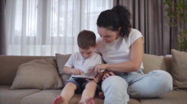 Sevimli bir aile, mutlu bir anne ve tatlı bir çocuk. Tablet bir cep telefonunun ekranına bakarlar. Çocuklu bir anne, evde kanepede oturur, akıllı telefon kahkahalarıyla oynar..