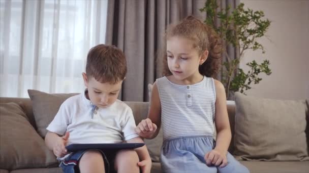 Linda familia e hija, cuñada una niñera con hermano pequeño, mirando la pantalla de una tableta en un teléfono celular, hija y su hermano están sentados en un sofá, jugando en casa en un teléfono inteligente . — Vídeo de stock