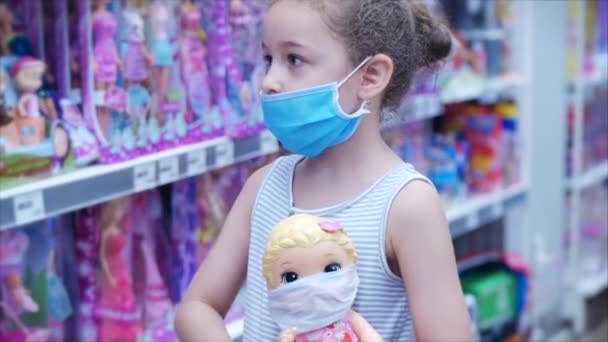 In de Store Een schattig klein meisje met een masker van virussen van de coronavirus epidemie kijkt naar de camera houdt haar pop in haar handen op de pop als een middel om het masker te beschermen tegen virussen, gemaskerd — Stockvideo