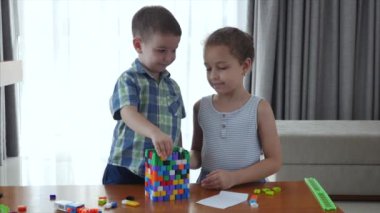 Mutlu bir aile, bir kız ve bir oğul, evde iki küçük çocuk bir masada oturup inşaatçıda oynuyorlar, bir kule inşa ediyorlar, oyuncu çocuklar birbirlerine yardım ediyorlar, oyuncaklarla eğleniyorlar.