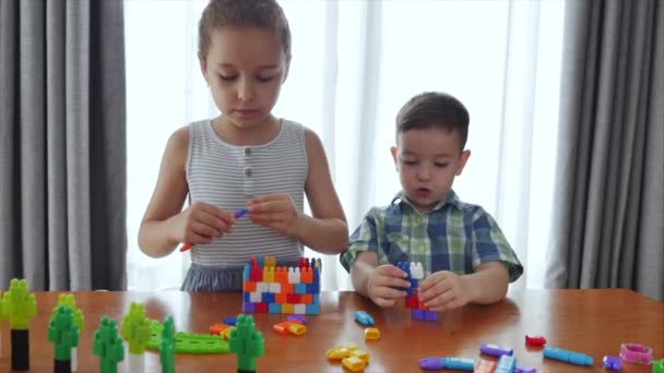 Famiglia felice, una figlia e un figlio, due bambini piccoli a casa si siedono a un tavolo e giocano nel costruttore, costruiscono una torre, i bambini giocherelloni si aiutano a vicenda, divertendosi a giocare con i giocattoli nel soggiorno. — Video Stock