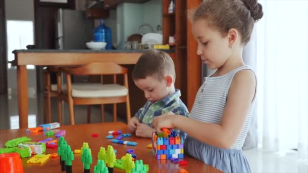 Familia feliz, una hija y un hijo, dos niños pequeños en casa se sientan en una mesa y juegan en el constructor, construyen una torre, los niños juguetones se ayudan unos a otros, se divierten jugando con juguetes en la vida — Vídeos de Stock