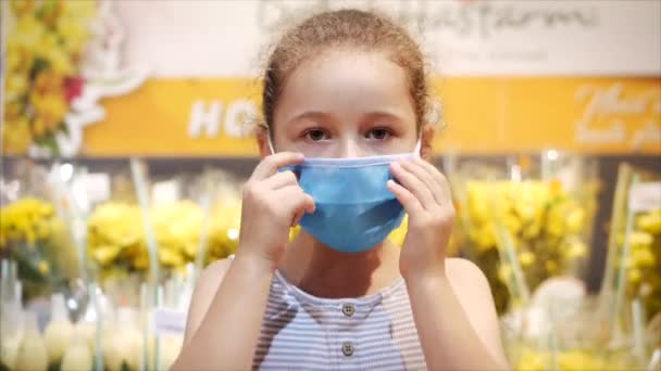 小さな女の子は黄色の花の背景にカメラを見てコロナウイルスやウイルスの流行のウイルスからマスクを修正します。コロンチン隔離された人々. — ストック動画