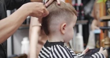 Ufaklık akıllı telefondan çizgi film izlerken usta saçını kesiyor. Covid-19 'un koruyucu maskeli stilisti küçük bir çocuk için şık bir saç stili yapıyor..