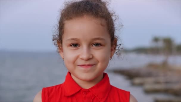 Grappig klein meisje glimlachend kind kijkt naar de camera op straat, een schattig kind staat op de asfaltweg tegen de achtergrond van straatbomen van palmbomen. — Stockvideo