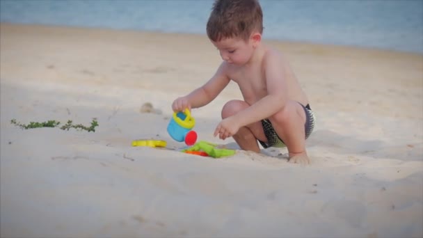 Niño feliz y despreocupado juega junto al mar, recoge la arena con una espátula, vierte agua de mar en una regadera, vierte arena. El pequeño juega junto al mar. Feliz infancia. . — Vídeos de Stock