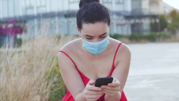 Ung kvinna i mask från coronavirus epidemin sitter på stranden nära havet i ensamhet, typer på sin smartphone, begreppet människor på självisolering från panik om den globala epidemin. — Stockvideo