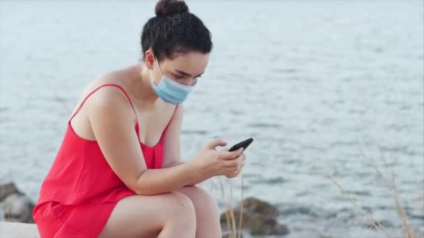 Young woman in a mask from the coronavirus epidemic sits on the shore near the sea in solitude,types on her smartphone,concept of people on self-isolation from panic about the global epidemic. — Stock Video