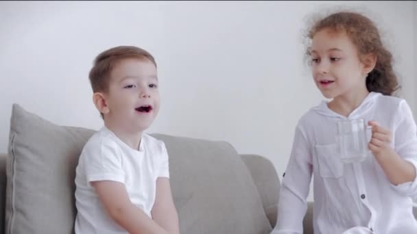 Filha uma irmã bonita, babá com um irmão mais novo estão sentados no sofá, amorosa irmã feliz cuida de seu irmão mais novo, diverte a criança, alimenta-lo mingau de arroz com peixes, ensina-lhe — Vídeo de Stock