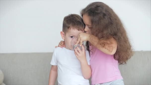 Mooie zus, een nanny met een jongere broer zitten op de bank, een liefhebbende gelukkige zus zorgt voor haar broertje, geeft een mok water, vermaakt het kind. — Stockvideo