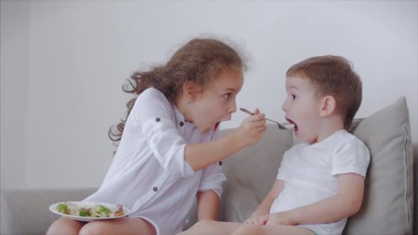 Daughter a pretty sister, nanny with a younger brother are sitting on the couch,loving happy sister takes care of her little brother, amuses the child,feeds him rice porridge with fish, teaches him — Stock Video