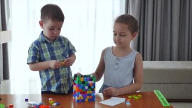 Mutlu bir aile, bir kız ve bir oğul, evde iki küçük çocuk bir masada oturup inşaatçıda oynuyorlar, bir kule inşa ediyorlar, oyuncu çocuklar birbirlerine yardım ediyorlar, oyuncaklarla eğleniyorlar.