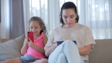 Mutlu bir aile, anne ve tatlı bir kız çocuğu, anne evde çocuklarla oynuyor rahatlamak için akıllı bir telefon kullanıyor kanepeye oturuyor, komik videolar izliyor, eğleniyor, aile hayatının tadını çıkarıyor.