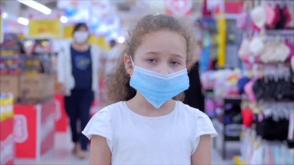 コロナウイルスやウイルスの流行からかわいい女の子は、パニックになって買い物をしているウイルスの仮面の人々の中にカメラを見ています。コロンチン人の隔離. — ストック動画