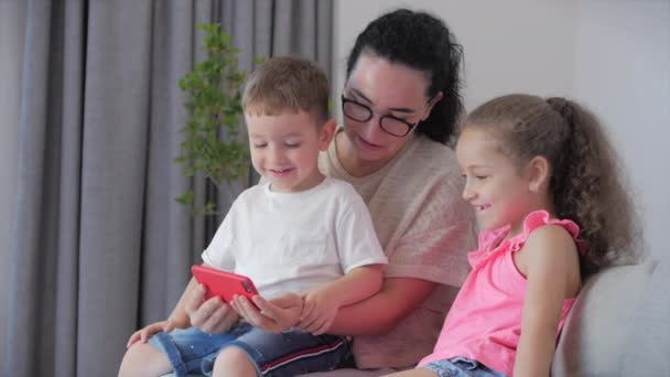 Familia feliz, mamá y lindos niños pequeños, madre jugando con los niños en casa relajante uso de un teléfono inteligente abrazo sentarse en el sofá hija e hijo risa, ver videos divertidos, divertirse, disfrutar de momentos de la vida familiar — Vídeo de stock