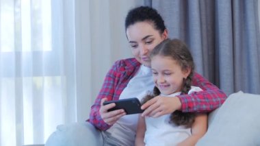 Mutlu bir aile, anne ve tatlı bir kız çocuğu, anne evde çocuklarla oynuyor rahatlamak için akıllı bir telefon kullanıyor kanepeye oturuyor, komik videolar izliyor, eğleniyor, aile hayatının tadını çıkarıyor.