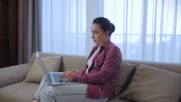 Joven mujer seria freelancer trabajando en una computadora portátil sentada en un sofá en casa, mujer de negocios escribiendo en una computadora portátil, chica enfocada usando una computadora para estudiar trabajo en línea en casa. Concepto de trabajo del empleado doméstico — Vídeos de Stock