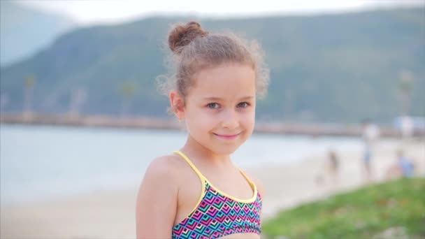Zabawna Happy Little Girl uśmiechnięta patrząc w kamerę na plaży w niebieskiej sukience z palmą, słodkie dziecko patrząc w aparat, uśmiechnięty i trochę nieśmiały. Dziecko, dzieci, emocje. — Wideo stockowe
