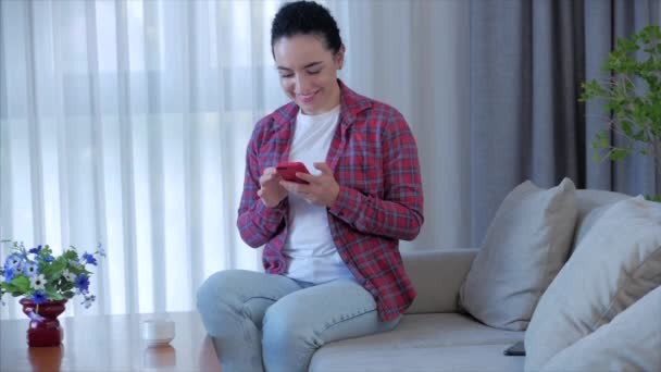 Serbest çalışan genç, ciddi bir kadın evdeki koltukta oturuyor, iş kadını evde oturup kahve içiyor ve evdeki online işleri öğrenmek için akıllı telefon çekleri yazıyor.. — Stok video